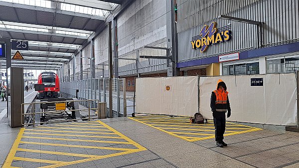In der rechten Bildhäfte verläuft diagonal ein Baustellenzaun als Absperrung. Davor steht jemand in orangefarbiger Jacke, links auf Gleis 25 ein roter Zug