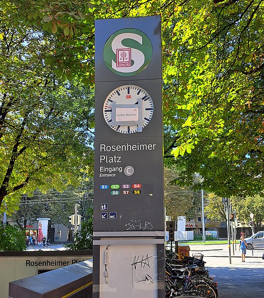 Eine graue Säle oder Stele, oben ein S-Bahn-Logo (grün, schlechter Zustand, illegaler Aufkleber), darunter eine Uhr mit DB-Logo, auf die ein Schild "Außer Betrieb", ebenfalls mit DB-Logo geklebt wurde. Darunter die Beschriftung "Rosenheimer Platz, Eingang C, Entrance" und eine nicht mehr ganz korrekte Liste von S-Bahn-Linien sowie Symbole für Treppe und WC. Darunter befindet sich eine leere Glasvitrine, die von Klebeband zusammengehalten wird.
Links sieht man den eigentlichen Zugang zum Untergeschoss, hinter der Stele abgegestellte Fahrräder und eine Straßenkreuzung. Über das Ensemble wölben sich Baumkronen.
