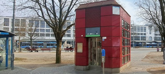 Dunkelrot verkleideter Aufzugzugang auf einer Platzfl&auml;che. Davor ein blauer MVV-Entwerter, sowie Bedientaste und Kontrollleuchte f&uuml;r den Aufzug. Im Hintergrund ein wei&szlig;er Geb&auml;uderiegel (etwa vier bis f&uuml;nf Stockwerke) mit zahlreichen Fenstern im Stil der 1970er/1980er-Jahre. Davor sieht man blaue Linienbusse. Links am Rand die Ecke eines MVG-Warteh&auml;uschens.