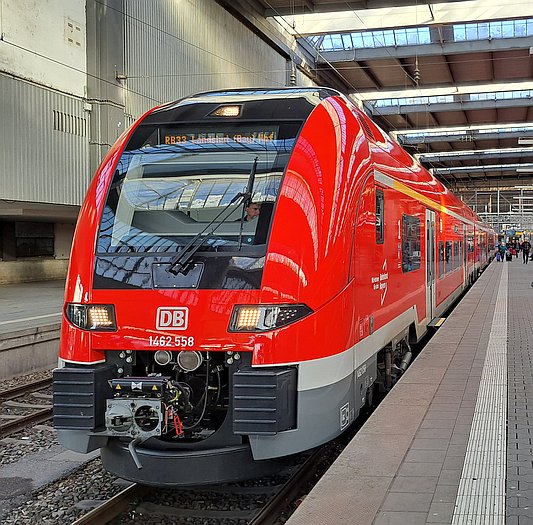 Ein roter Nahverkehrszug der Deutschen Bahn steht an einem Bahnsteig im Münchner Hauptbahnhof. Weiter hinten auf dem Bahnsteig sind Menschen zu erkennen. Es handelt sich um einen Zug des Typs Desiro HC, unter dem DB-Logo steht die Zugnummer 1462 558. Darunter sind die Kupplung und weitere technische Einrichtungen zu erkennen. Der Zug scheint neu zu sein, da sein roter Lack gl&azml;nzt. Der Zug ist von schrüg vorne fotografiert mit Fahrtrichtung aus dem Hauptbahnhof hinaus. In der Zielanzeige ist, etwas durch Reflexionen gestört, 'RB33 Landshut (Bay) Hbf' zu lesen.  Links ist die Seitenwand der Bahnhofshalle an Bahnsteig 26 zu sehen und oben die Hallendecke.