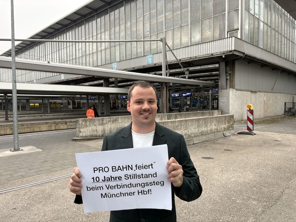 Eine Person steht vor dem Hallenende des Münchner Hauptbahnhofs im Bereich der Unterführung Paul-Heyse-Straße in der Nähe des Bahnsteigs von Gleis 11 und zeigt ein Schild mit der Aufschrift "PRO BAHN 'feiert' 10 Jahre Stillstand beim Verbindungssteg Münchner Hbf!