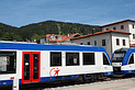 Fahrzeuge der Bayerischen Regiobahn im Bahnhof Schliersee. Im Hintergrund rechts ein Teil des Bahnhofsgebädes, in der Ferne bewaldete Berge.