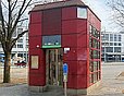 Dunkelrot verkleideter Aufzugzugang. Im Hintergrund ein wei&szlig;er Geb&auml;uderiegel (etwa vier bis f&uuml;nf Stockwerke) mit zahlreichen Fenstern im Stil der 1970er/1980er-Jahre.