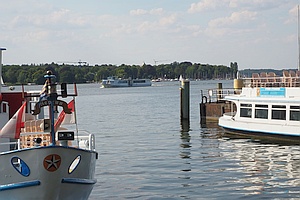 Im Vordergrund Teile zweier Bug voraus am Ufer 
liegenden Schiffe, dazwischen im Hintergrund die Wannseefähre