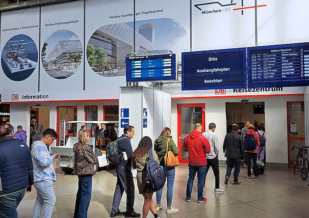 mehrere Menschen stehen in einer Schlange, teilweise mit Gepäck, im Hintergrund Eingang mit Aufschrift "Reisezentrum" darüber Anzeigmonitore für Zugabfahrten, einer mit Aufschrift "Bitte Aushangfahrplan beachten",auf der Wand dahinter Werbung für 2 Stammstrecke und Neubau Empfangsgebäde des Hauptbahnhofs und Starnberger Flügelbahnhof