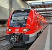 Ein roter Nahverkehrszug der Deutschen Bahn steht an einem Bahnsteig im Münchner Hauptbahnhof. Es handelt sich um einen Zug des Typs Desiro HC, unten sind die Kupplung und weitere technische Einrichtungen zu erkennen. Der Zug scheint neu zu sein, da sein roter Lack glänzt. Der Zug ist von schrüg vorne fotografiert mit Fahrtrichtung aus dem Hauptbahnhof hinaus. Links ist die Seitenwand der Bahnhofshalle an Bahnsteig 26 zu sehen und oben die Hallendecke.