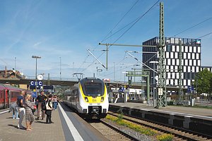 Bahnsteig mit einfahrendem Zug (Baureihe 442) in den Farben wei&szlig;, gelb und schwarz. Im Hintergrund eine Br&uuml;cke und rechts ein Hochhaus.