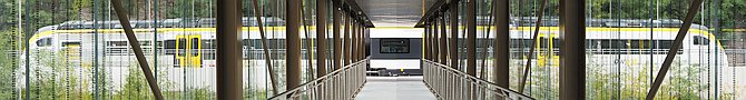 Blick durch einen &uuml;berdachten Gang, rechts und links leicht gemusterte Glasw&auml;nde und braune Stahlträger. Im Hintergrund ein wei&szlig;-gelber Zug. Das Bild ist eine Montage aus zwei Fotos.