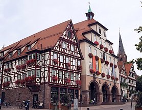 In der Mitte große verputztes Haus mit kleinem Turm, rechts und links Fachwerkh&auml;user, im Hintergrund ein Kirchturm.