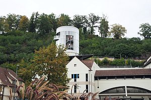 In der Mitte oben ein runder Turm, in dem eine Treppe und Menschen erkennbar sind. Im Hintergrund ein bewachsener Hang. Unterhalb des Turms weitere Geb&auml;ude und rechts unten eine Art Torbogen.