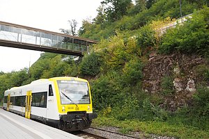 Im Vordergrund ein gelb-wei&szlig;er Dieseltriebwagen (Regioshuttle) am Bahnsteig. Dar&uuml;ber eine Br&uuml;cke mit gl&auml;sernen Seitenw&auml;nden. Rechts oben, kaum erkennbar, ein weiterer Zug am oberen Bahnsteig.
