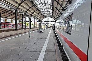 ICE steht am Bahnsteig in einer historischen Bahnhofshalle. Blick Richtung Hallenende. In etwas Entfernung erkennt man einige Menschen. Ein paar Gleise weiter links ein Regionalzug.