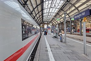 ICE steht am Bahnsteig in einer historischen Bahnhofshalle. Blick Richtung Prellbock. Auf dem Bahnsteig ein Schild 'Wiesbaden Hbf' und einige Menschen. Ein paar Gleise weiter rechts ein Regionalzug.