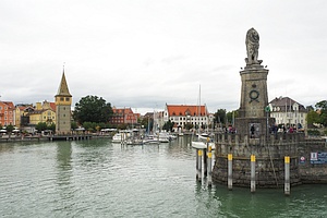 Wasserfläche mit einer auf einem Vorsprung stehenden Löwenstatue, im Hintergrund ein historisch wirkender Turm, Gebäude und festgemachte Boote