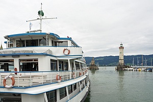 im Vordergrund links ein Passagierschiff (Teilansicht), rechts im Mittelgrund ein Leuchtturm, im Hintergrund Berge