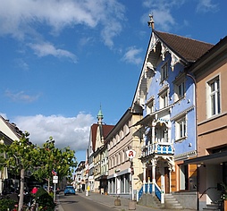 Straßenzug mit historischen Häusern auf der rechten Seite, im Vordergund ein hellblau gefärbtes mit verziertem Giebel, Balkon, Treppenzugang und einem Apothekenschild