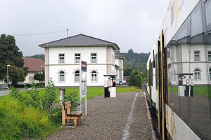 rechts Außeneite eines Zuges, Mitte unten Bahnsteig mit gekiester Oberfläche und einer Holzbank, in Hintergrund großes Gebäude mit Aufschrift 'Krauchenwies'