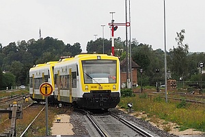 gelb-weißer Zug, regnerisches Wetter, im Hintergrund ein Formsignal, rechts eine offene Halle mit abgestellten LKW