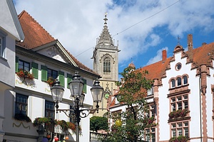zwei historische Gebäude in unterschiedlichen Baustil (jeweils Teilansicht), im Vordergrund eine Laterne, im Hintergrund ein Kirchturm