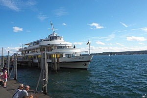 ein Passagierschiff mit Namen Stuttgart an einer Anlegestelle, unten links sieht man im Vordergrund Leute am Ufer sitzen, in der Fern Segelboote und das gegenüberliegende Ufer