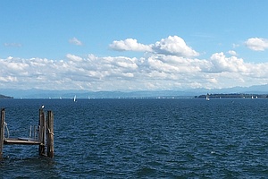 ein See, links ragt ein Steg ins Bild, in der Ferne Segelboote und ganz hinten Berge