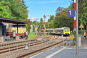 Blick auf eine Bahnhofseinfahrt, links ein Bahnsteig und eine Diesellok, in der Mitte Formsignale, auf dem rechten Gleis ein einfahrender Zug in gelb-weißer Farbgebung