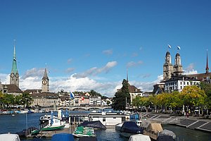 Fluss mit Liegeplätzen für Boote, rechts und links Bebauung mit herausragenden Kirchtürmen, rechts die zwei Türme des Zürcher Großmünsters mit blau-weißen Flaggen