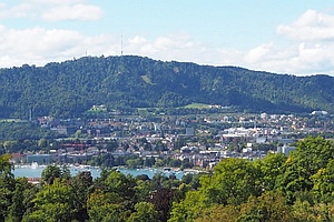 Panorama mit städtischer Bebauung und einem Stück Seefläche, im Vordergrund Bäume, im Hintergrund ein Höhenzug