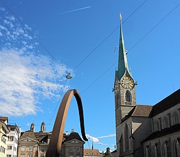 rechts eine Kirche mit spitzem Turm, im Vordergrund der geschwungene Einlauf eines Brunnens, im Hintergrund weitere Gebäude über denen man die Turmspitzen des Zürcher Großmünsters sieht