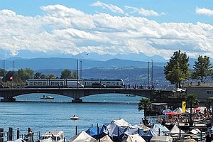 Blick auf Fluss mit Brücke, über die eine Tram fährt, im Vordergrund Liegeplätze mit abgedeckten Booten, im Hintergrund hohe Berge