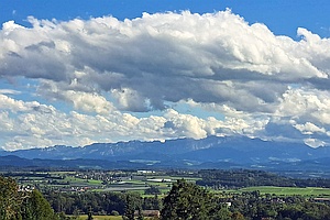 hinter einer locker besiedelten Ebene sieht man hohe Berge, darüber eindrucksvolle Wolkengebilde