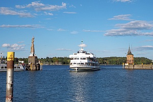 weißes Passagierschiff 'Stuttgart' färt in Hafen hinein, auf der linken Hafenmole steht eine große Frauenfigur ('Imperia'), rechts ein kleines Gebäude