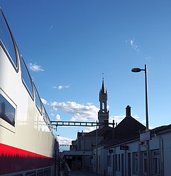 links Teilansicht eines Doppelstockzuges in weiß mit rotem Zierstreifen, rechts Gebäude mit schmalem spitzen Turm