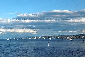 Blick auf See mit Segelbooten und kleinerem Passagierschiff, im Hintergrund bebautes Ufer, am Himmel viele Wolken